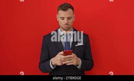 A professional-looking man in a suit interacts with a smartphone against an isolated red background. Stock Photo