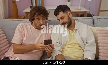 Two men sit closely together on a comfortable couch indoors, focused on a smartphone screen, indicating the warmth and bond of their relationship in a Stock Photo