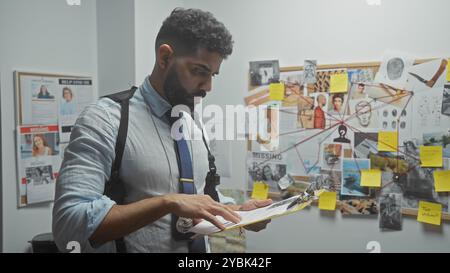 A focused hispanic man with a beard examines a clipboard in a detective's office surrounded by investigation evidence. Stock Photo