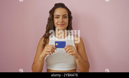 Young woman holding credit card in front of isolated pink background, smiling confidently and wearing white crop top Stock Photo