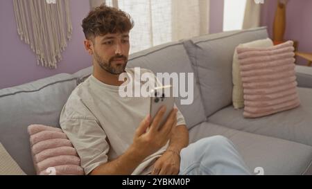 Young man sitting on a couch at home, looking at his smartphone in a modern living room. Stock Photo