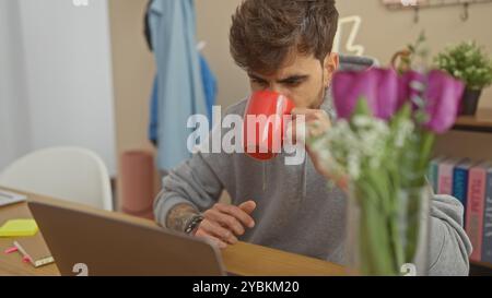 A young hispanic man sips coffee indoors at a home office with a laptop, books, and flowers in the background. Stock Photo
