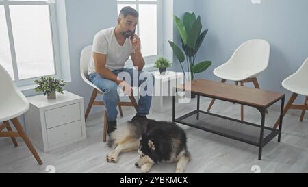 A young hispanic man in casual attire talks on the phone beside his husky dog in a bright veterinary waiting room. Stock Photo