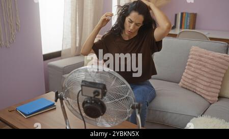 Young woman feeling hot while sitting in living room, trying to cool down with a fan on a summer day. Stock Photo
