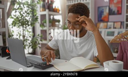 Young man using a calculator in a home decor store, surrounded by indoor plants and decorations, focused on work with a laptop and a notebook. Stock Photo
