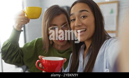Two smiling women taking a selfie in an office, holding colorful coffee mugs and enjoying a casual work moment with a cheerful and friendly atmosphere Stock Photo
