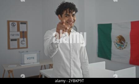 Smiling young hispanic man pointing, wearing a white shirt in a voting room with a mexican flag in the background Stock Photo