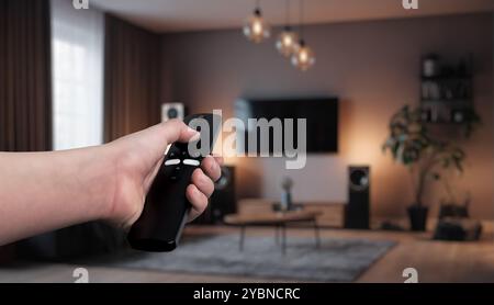 Hand holding a remote control, adjusting smart home devices in a modern living room. Concept of home automation, controlling lights, curtains, and sma Stock Photo