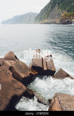 Concrete breakwater blocks along Seixal's coast with a backdrop of lush cliffs and the vast Atlantic Ocean Stock Photo