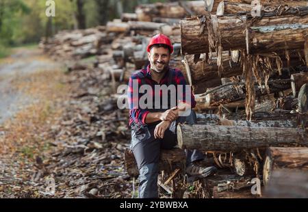 Handsome man lumberjack with helmet sitting on tree trunk stacked in forest Stock Photo