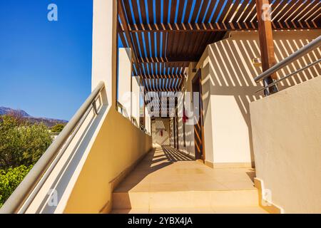 Sunny corridor hotel with shadows from wooden pergola on tiled floor leading to doors. Crete. Greece. Stock Photo