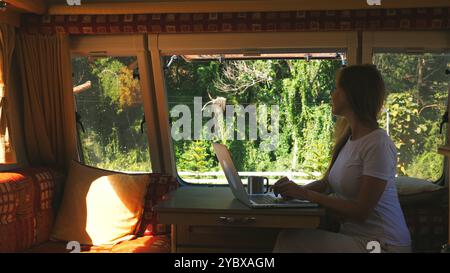 Young woman working on laptop inside cozy RV, looking out window at Stock Photo