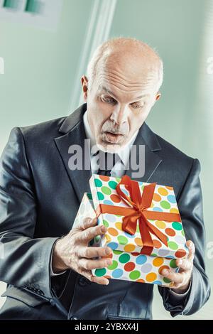 Older businessman in a suit is opening a colorful gift box with a surprised expression on his face. The image evokes feelings of joy and anticipation, Stock Photo