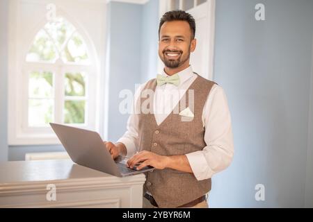 A man in a vest and bow tie smiles while working on a laptop Stock Photo