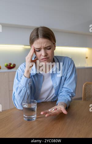 Struggling grimacing woman with headache drinks antidepressants in kitchen. Female mental problems Stock Photo