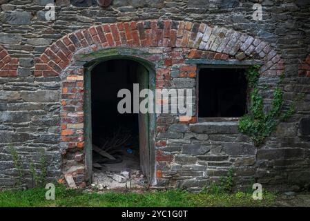 The wall of an abandoned, industrial building shows crumbling stone and brick, flanked by climbing ivy, evoking a sense of decay and neglect. Photo ta Stock Photo