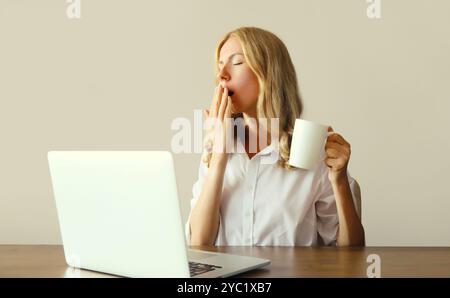 Tired overworked sleeping woman employee yawns, drinks coffee while working with laptop sitting at desk in office in morning Stock Photo