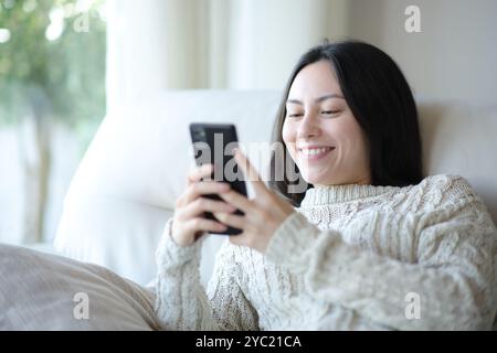 Woman in sweater checking cell phone sitting on a sofa in winter at home Stock Photo