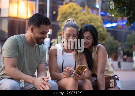 Three friends laughing and using a smartphone outdoors in the city, showcasing the joy of friendship and technology in the summer Stock Photo