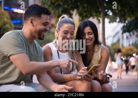 Three happy friends sitting outdoors on a bench, using a smartphone and sharing a laugh together, capturing the essence of togetherness and youthfulness in the digital age Stock Photo