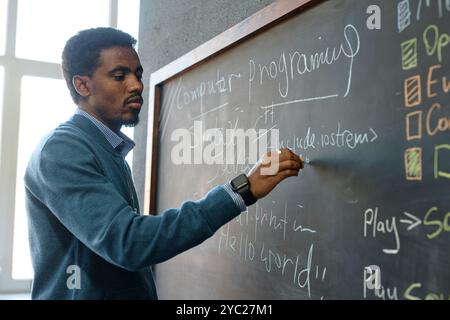 Side view portrait of young African American male teacher writing on blackboard in school classroom explaining basics of IT programing copy space Stock Photo