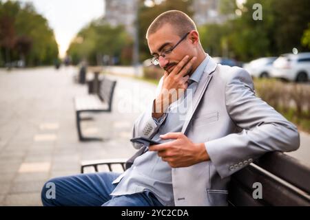 Portrait of pensive businessman who is messaging on phone. Man sitting on bench in the city and using smartphone. Stock Photo