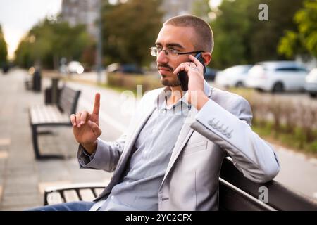 Portrait of pensive businessman who is talking on phone. Man sitting on bench in the city and talking on phone. Stock Photo