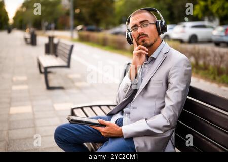 Portrait of thoughtful businessman who is sitting on bench in the city and using digital tablet. Stock Photo