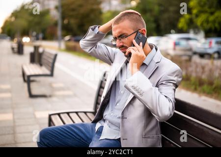 Portrait of worried businessman who is talking on phone. Man sitting on bench in the city and talking on phone. Stock Photo