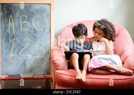Two children sitting together on a pink chair, using a tablet, w Stock Photo