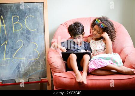 Two children sitting on a pink chair, using a tablet together, w Stock Photo