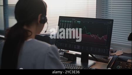 Modern office of International stock exchange company: Back view of female specialist in headset working on computer, communicating with client. PC screen shows equity market and shares in real-time. Stock Photo