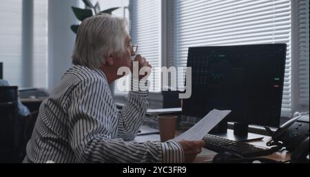 Tired senior financial specialist analyzes papers and documents, monitors stock market and shares on computer. Professional trader works in the modern office of international stock exchange company. Stock Photo