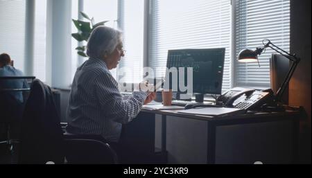 In modern office: Senior financial trader or businessman works in international stock exchange company. Specialist uses mobile phone, analyzes and monitors stock market and shares on personal computer Stock Photo