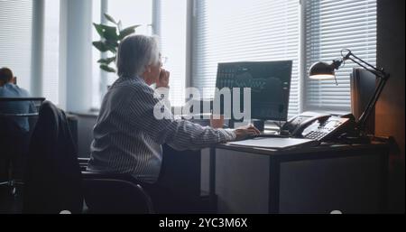 In modern office: Senior financial trader or businessman works in international stock exchange company. Specialist uses mobile phone, analyzes and monitors stock market and shares on personal computer Stock Photo