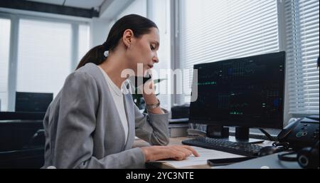 Female financial analyst discusses strategy of project with colleague using mobile phone. Specialist from marketing department checks data in documents with parameters displayed on PC monitor. Stock Photo