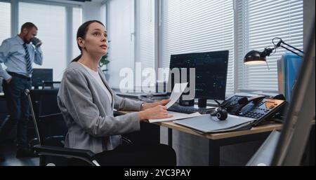 Workplace of managers of large financial company: Adult, beautiful female analyst checking data in paper documents for changes in foreign exchange rates. Monitoring stock and equity markets concept. Stock Photo