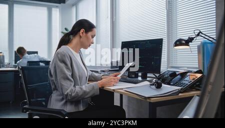 Workplace of managers of large financial company: Adult, beautiful female analyst checking data in paper documents for changes in foreign exchange rates. Monitoring stock and equity markets concept. Stock Photo