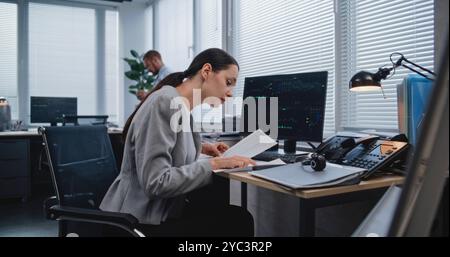 Workplace of managers of large financial company: Adult, beautiful female analyst checking data in paper documents for changes in foreign exchange rates. Monitoring stock and equity markets concept. Stock Photo