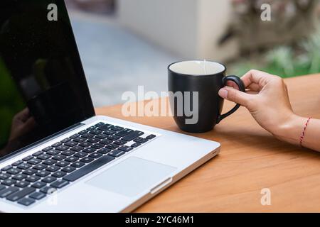 A person holding a black coffee mug while sitting at a wooden table with a laptop open. The laptop screen is dark, and the background features a blurr Stock Photo