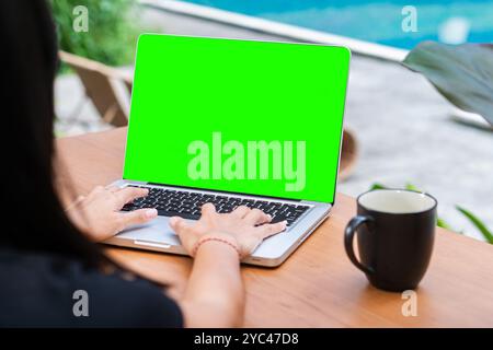 A person typing on a laptop with a green screen display. A black coffee mug is placed beside the laptop on a wooden table. The background is blurred, Stock Photo