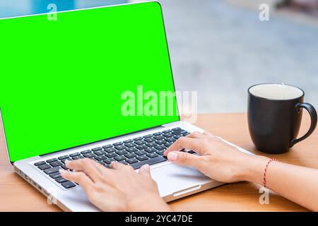 A person typing on a laptop with a green screen display. A black coffee mug is placed beside the laptop on a wooden table. The background is blurred, Stock Photo
