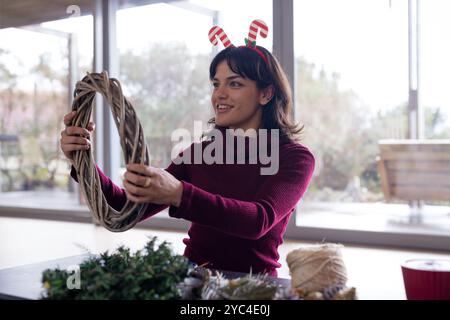Woman crafting Christmas wreath at home, wearing festive candy cane headband Stock Photo