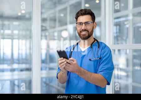 Smiling doctor in blue scrubs uses smartphone with stethoscope around neck in bright hospital. Engaging healthcare professional conveys trust, technology, modern medical communication. Stock Photo