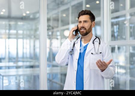 Confident male doctor in white coat converses on phone in modern office. Stethoscope around neck emphasizes medical expertise. Bright setting reflects professional atmosphere and communication. Stock Photo