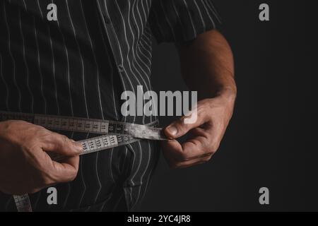 Man with big belly measures his waist with measuring tape on black background, closeup Stock Photo