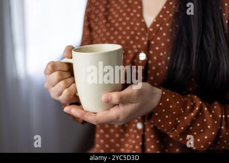 Close-up of asian woman holding mug, displaying comfort, warmth, and relaxation. Hands cradling neutral-colored cup, wearing red polka dot blouse, sug Stock Photo