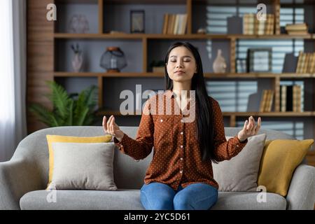 Asian woman practicing meditation on sofa, eyes closed, embracing calm and mindfulness. Dressed casually, sitting in relaxed pose. Living room setting Stock Photo