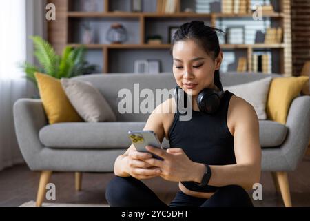 Asian woman in sportswear performs exercises, watches online fitness classes on phone, headphones around neck, in comfy home setting. Engaged in healt Stock Photo