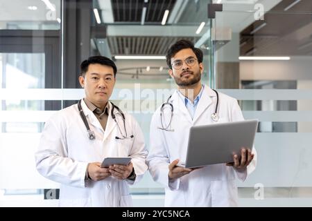 Doctors in white coats collaborating in modern office, using tablet and laptop for healthcare discussions. Stethoscopes visible, highlighting medical Stock Photo
